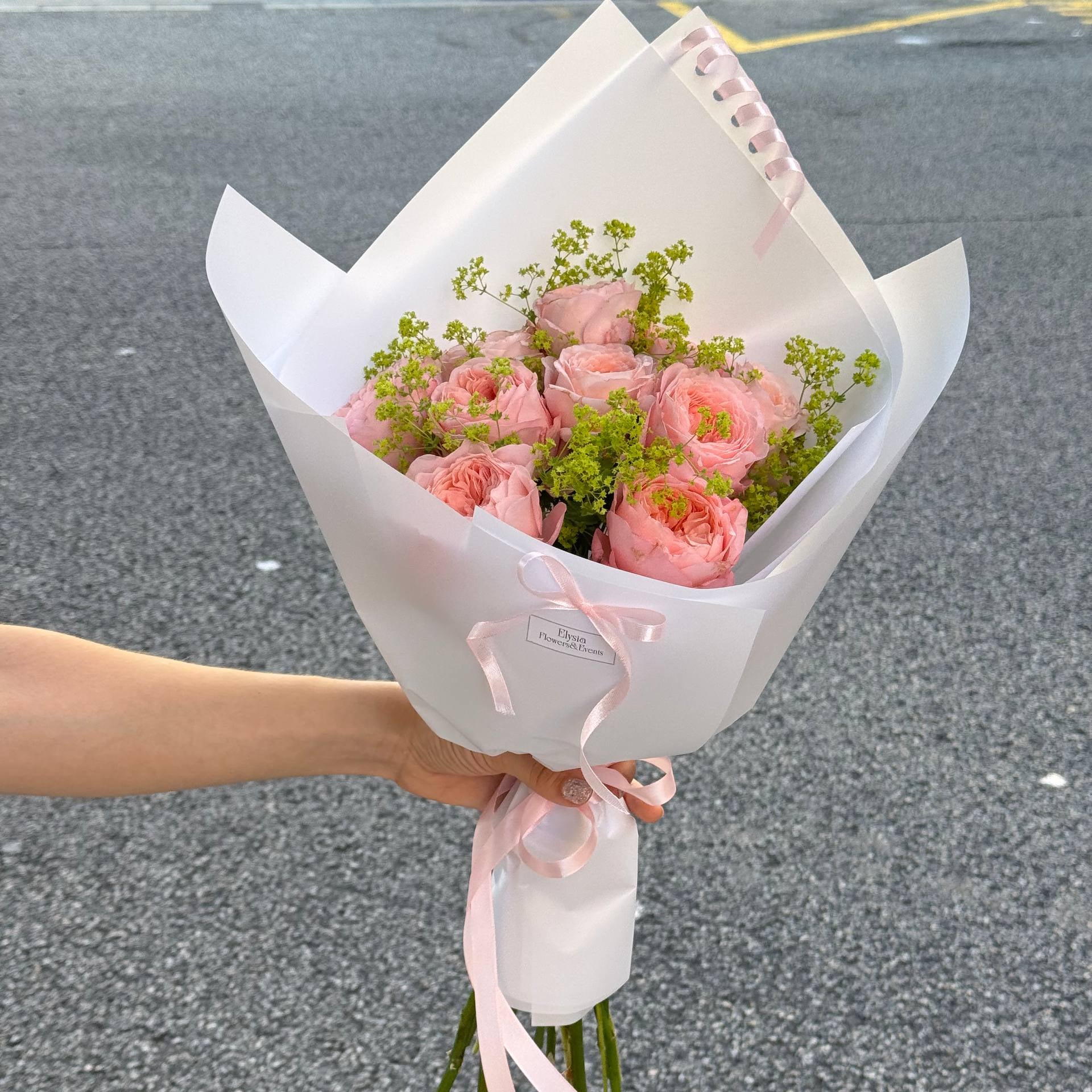 Bouquet of pink flowers wrapped in white paper with a pink ribbon held by a person on a gray pavement. Bouquet of pink flowers wrapped in white paper with a pink ribbon held by a person on a gray pavement.
