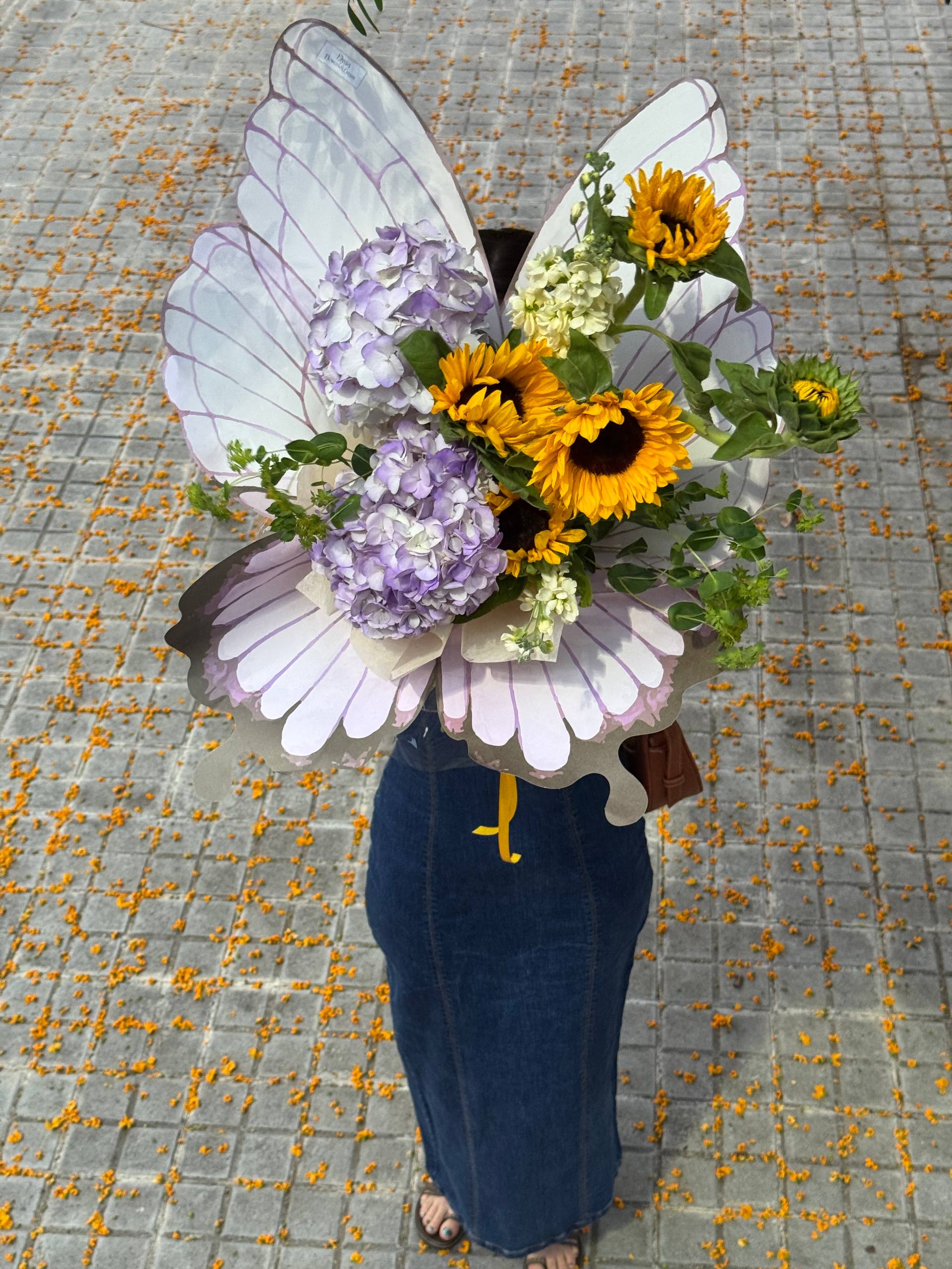 Person holding a bouquet shaped like butterfly wings with flowers on a textured ground. Persona sosteniendo un ramo con forma de alas de mariposa con flores en un suelo texturizado.
