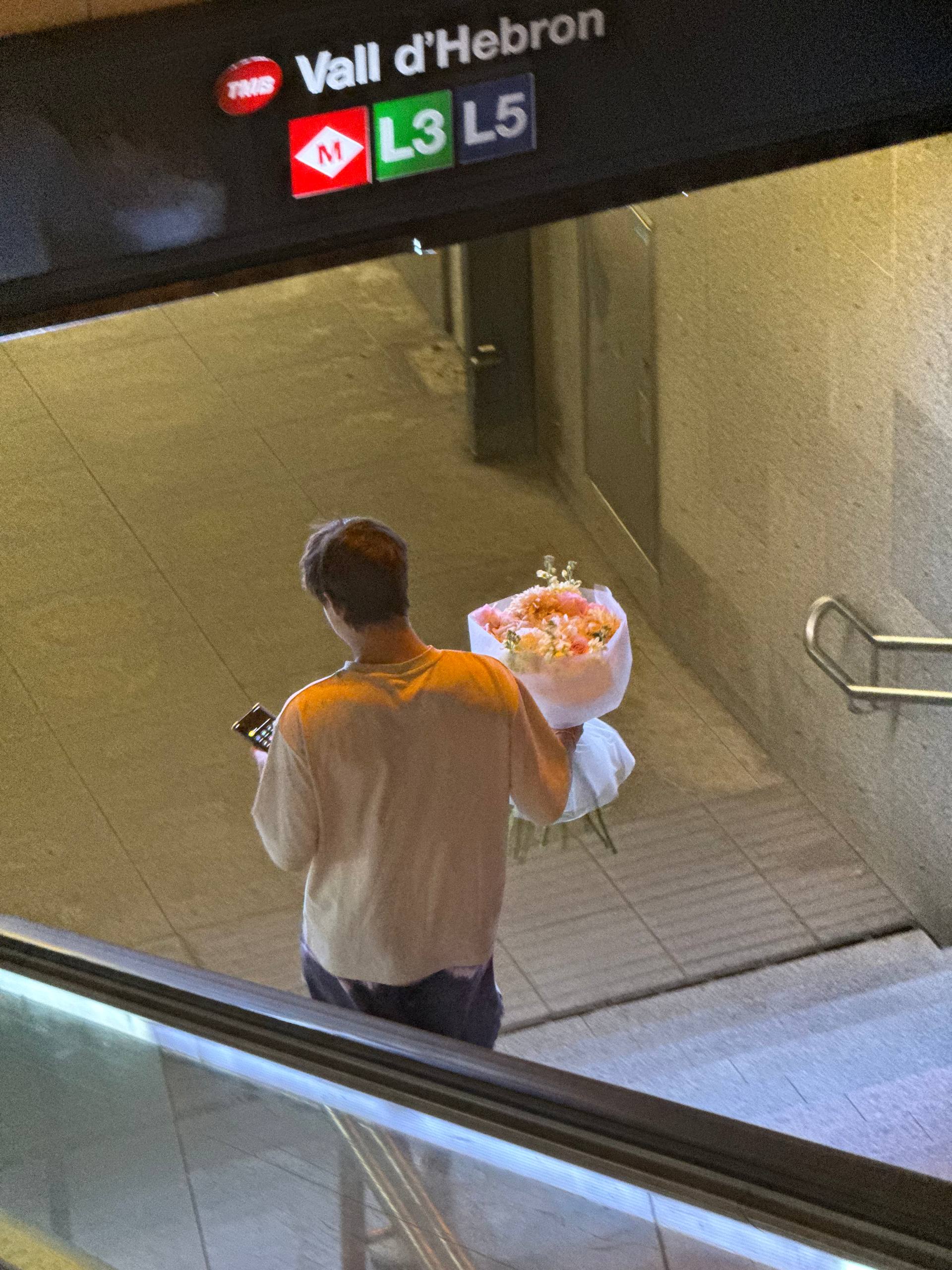 Person holding flowers and a phone in a metro station with a sign above. Persona sosteniendo flores y un teléfono en una estación de metro con un cartel arriba.