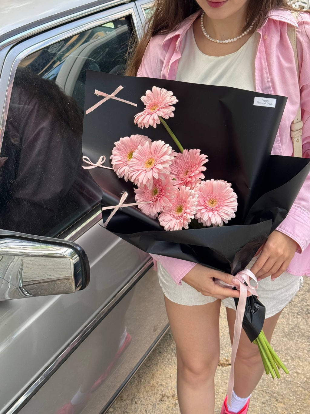 Person holding a bouquet of pink flowers in front of a car Persona sosteniendo un ramo de flores rosadas frente a un automóvil